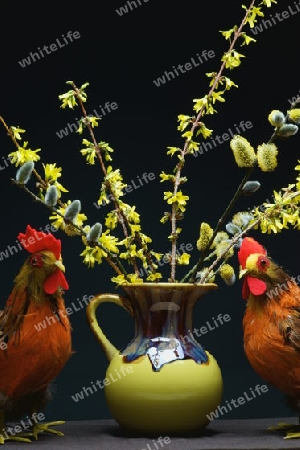 Ein Hahn und ein Huhn als Dekoration. In der Mitte eine gelbe Vase mit einem Strauss Weide und Forsythie.