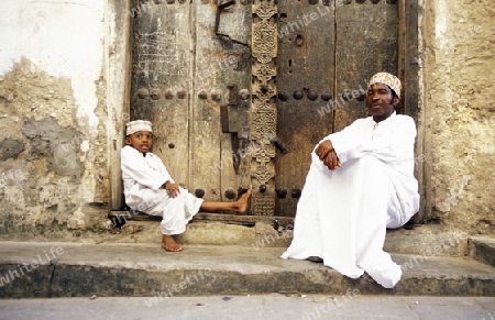  Ein Mann aus Zanzibar in traditionelle Kleidung steht an einer alten Holztuer inmitten der Altstadt Stone Town der Hauptstadt Zanzibar Town auf der Insel Zanzibar welche zu Tansania gehoert.     
