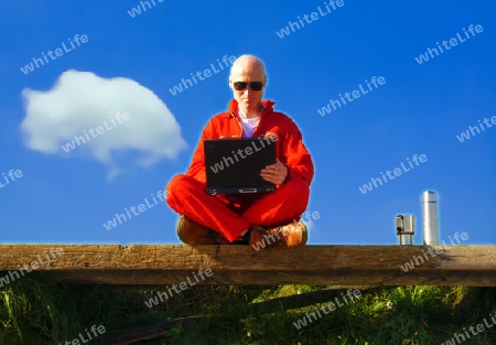 Man in red suite sitting on a bench at the beach under a blue sky working with his laptop - Mann in rotem Overall sitzt auf einer Bank am Strand und arbeitet mit dem Notebook