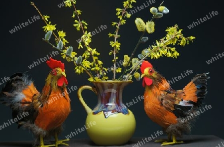 Ein Hahn und ein Huhn als Dekoration. In der Mitte eine gelbe Vase mit einem Strauss Weide und Forsythie.