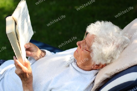 Alte Frau liegt im Garten auf Liege und liest Buch