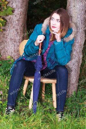 Junge Frau mit Regenschirm in sehr modischer Kleidung sitzt im Wald