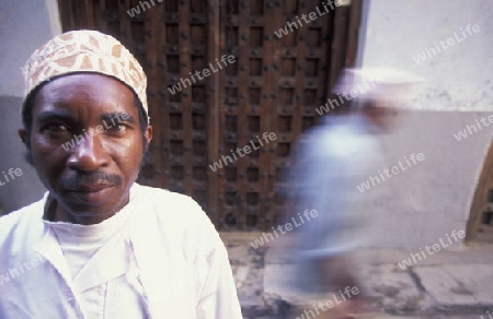  Ein Mann aus Zanzibar in traditionelle Kleidung steht an einer alten Holztuer inmitten der Altstadt Stone Town der Hauptstadt Zanzibar Town auf der Insel Zanzibar welche zu Tansania gehoert.     