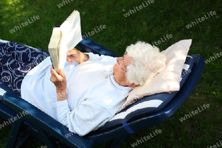 Alte Frau liegt im Garten auf Liege und liest Buch