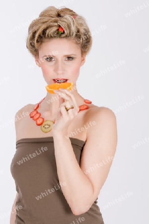 Attraktive blonde junge Frau mit Erdbeere-Kiwi- Kette isst eine Orange / Attraktive blonde junge Frau mit Erdbeere-Kiwi- Kette isst eine Orange